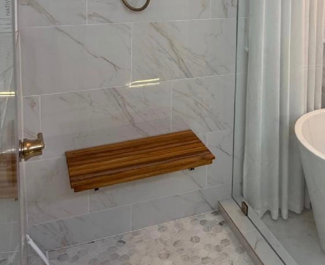 A wall-mounted folding teak wood shower bench with a rich wood grain finish, installed on a light gray marble-tiled bathroom wall next to a glass shower door and white bathtub.