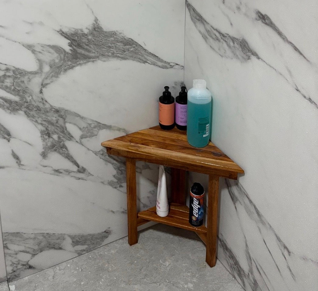 A triangular wooden corner shelf stool placed in a marble-tiled bathroom corner, holding several toiletry bottles.