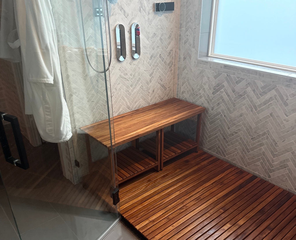 A walk-in shower featuring a wooden slatted teak mat with a matching rectangular freestanding wooden teak bench. The bench has two slatted shelves underneath. The shower has white and gray tile walls 