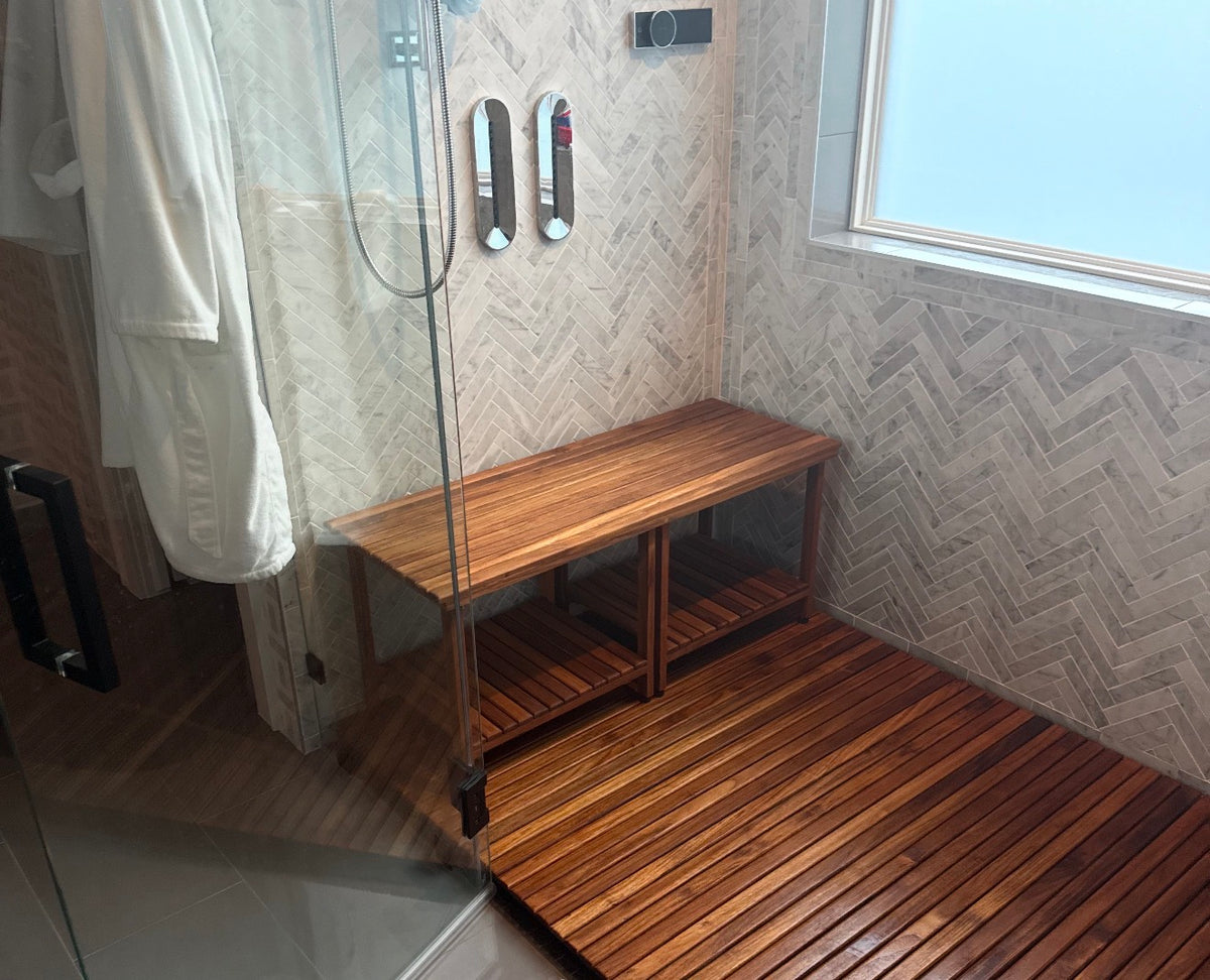 A walk-in shower featuring a wooden slatted teak mat with a matching rectangular freestanding wooden teak bench. The bench has two slatted shelves underneath. The shower has white and gray tile walls 