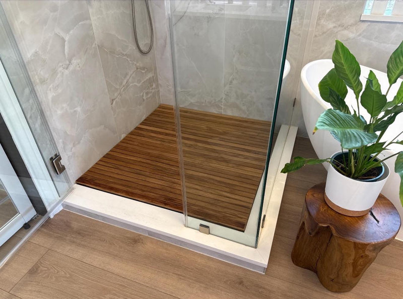 A modern walk-in shower enclosure with a brown wood-look slatted shower floor and clear glass panels, shown in a bathroom next to a white tub and a potted green plant on a wooden stool.