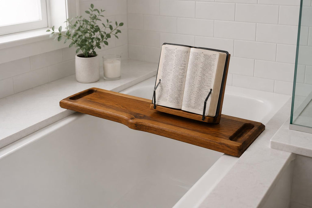 A wooden bath caddy resting across a white bathtub, holding an open book or Bible on a small black stand. The scene is set in a bright, modern bathroom with white subway tiles, a window, and a small potted plant.