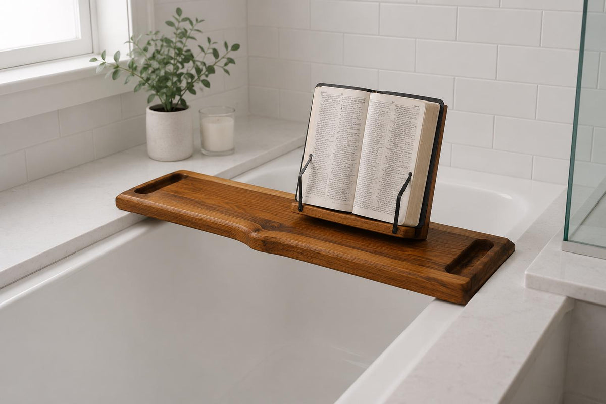 A wooden bath caddy resting across a white bathtub, holding an open book or Bible on a small black stand. The scene is set in a bright, modern bathroom with white subway tiles, a window, and a small potted plant.